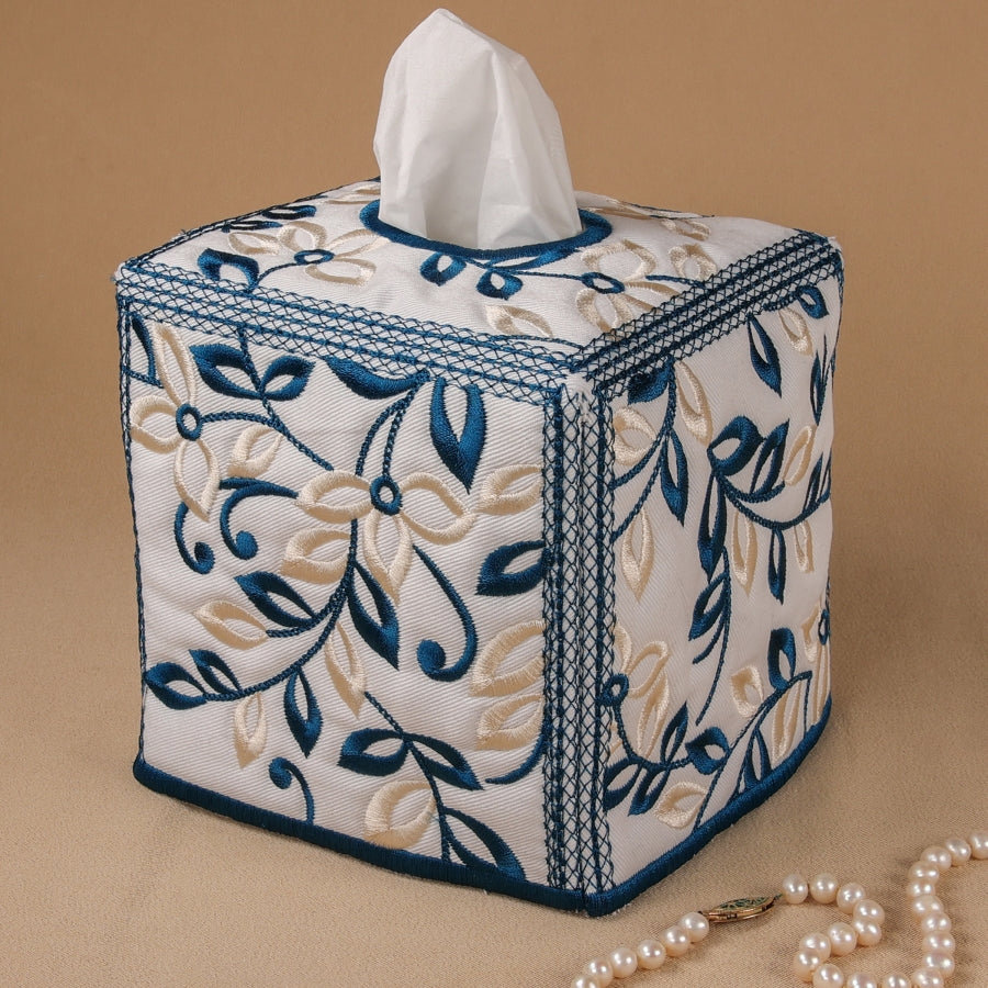 A square tissue box cover with a floral embroidery design in blue and beige, featuring a satin outline around the panels.