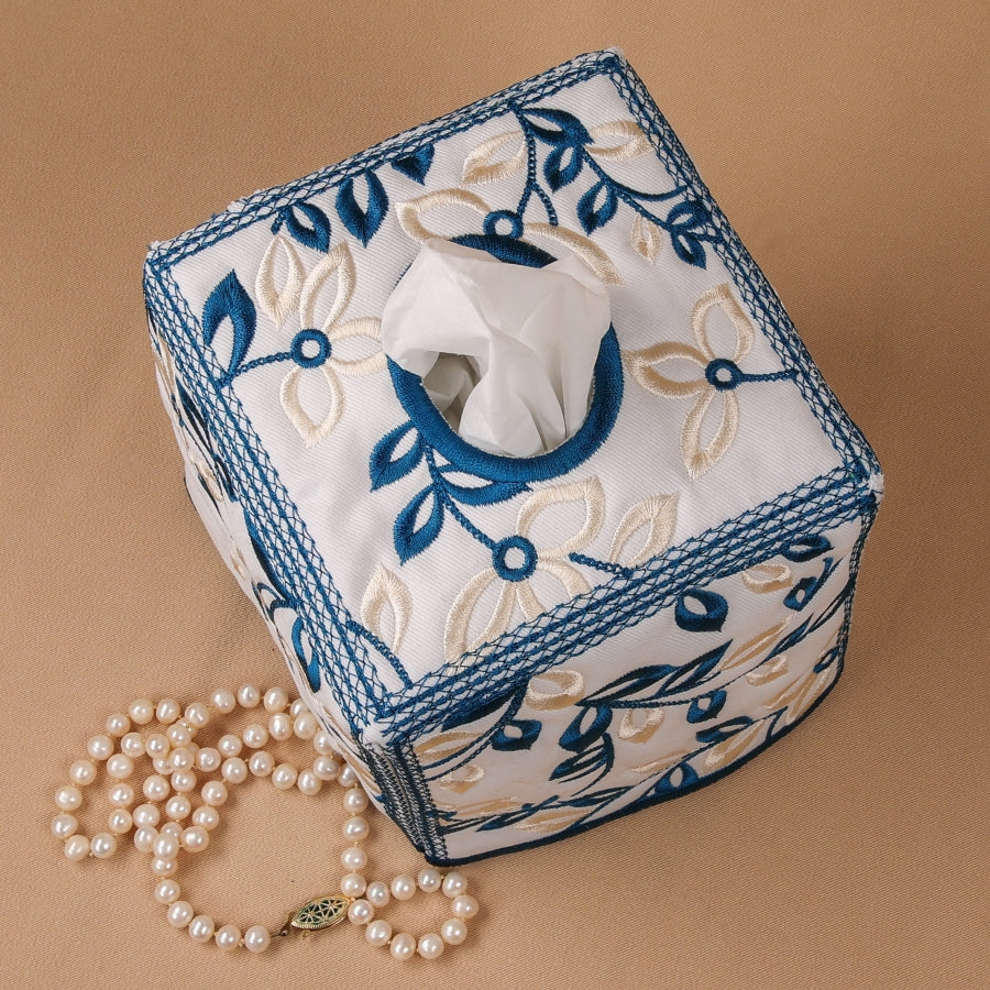 A square tissue box cover with a floral embroidery design in blue and beige, featuring a satin outline around the panels.