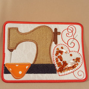 A square fabric patch featuring an applique design of a sewing machine, an orange sewing foot, and a heart-shaped sequin embellishment on a white background with a red border, set against a beige fabric backdrop.
