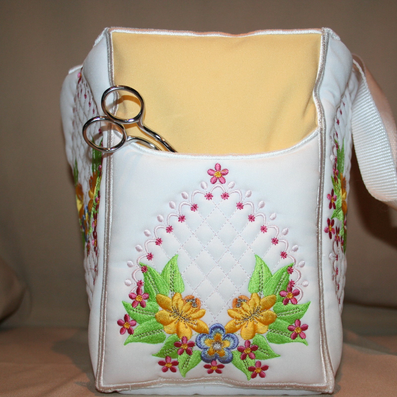 White tote bag with floral embroidery containing a bottle of lotion on a beige surface.