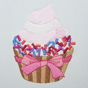 Embroidered design of a cupcake with pink frosting, sprinkles, and a bow on a light gray background.