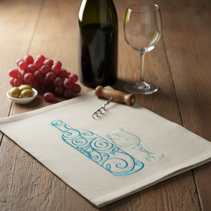 A beige kitchen towel with embroidered blue wine bottle and glass designs, styled on a wooden table with a wine bottle, wine glass, grapes, and olives by astitchahalf.net