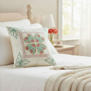 A decorative square throw pillow with a four-leaf clover motif in green and pink floral print, resting on a white bed in a softly lit bedroom.