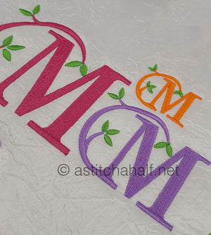 Embroidered letters 'MOM' in pink, orange, and purple with green leaves on a textured white background.