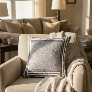 Beige sofa with a textured gray pillow and knitted blanket, next to a small table with a plant.
