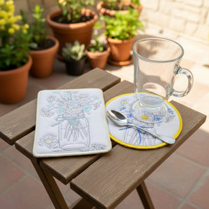 Two embroidered coasters with floral designs on a beige surface, one with a glass and spoon.