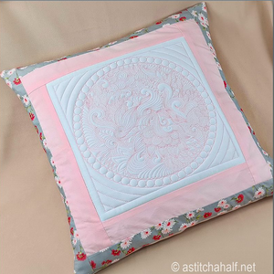 A square decorative pillow with a floral border and a central light blue quilted patch featuring pink embroidered details, displayed on a beige fabric background by astitchahalf.net