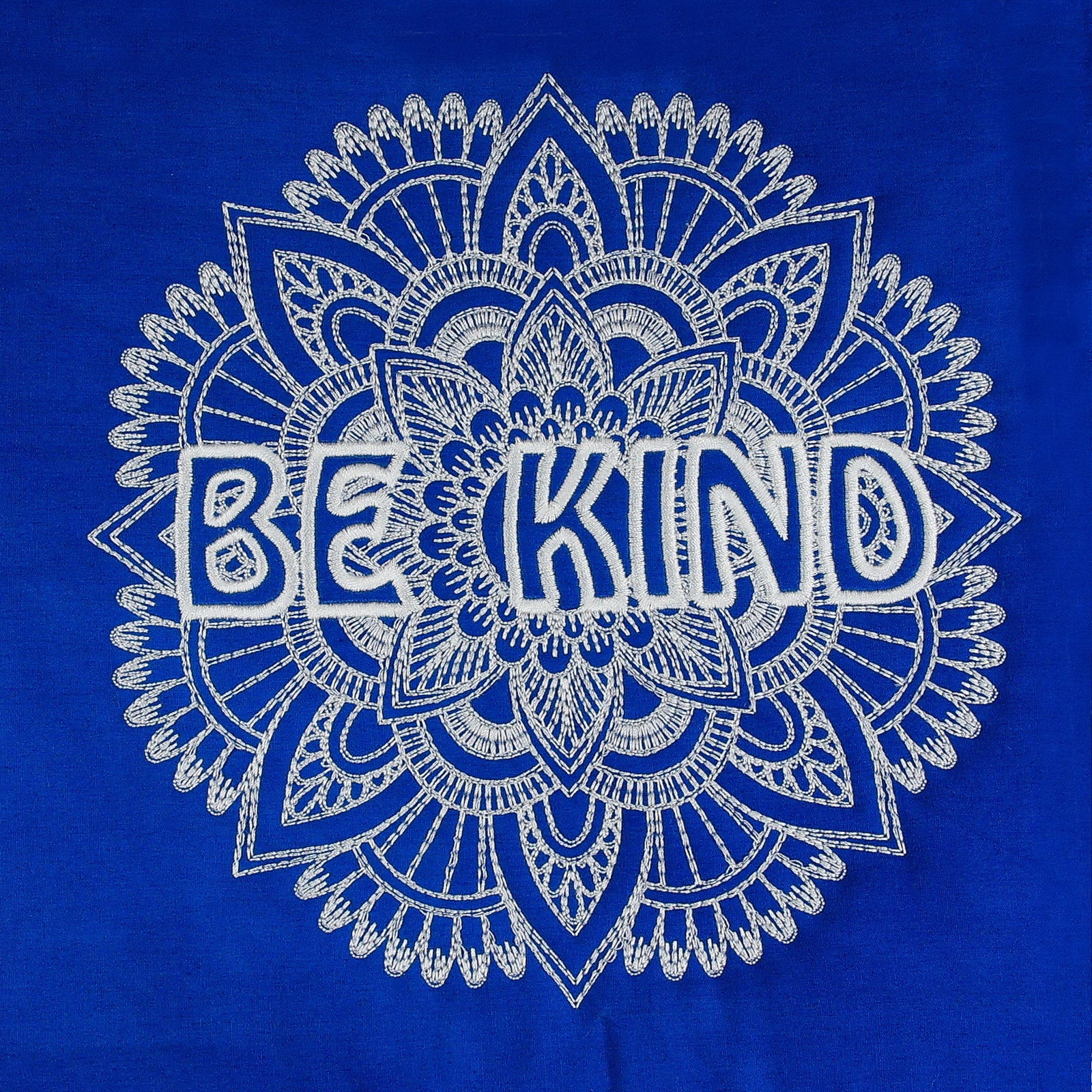 Blue tote bag with 'BE KIND' design hanging on a hook against a white wall.