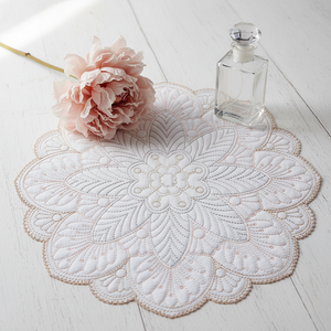 Embroidered white doily with a pink flower and clear glass container on a light wooden surface