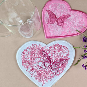 Embroidered heart-shaped trivet with a butterfly design, displayed alongside a mug and decorative flowers.