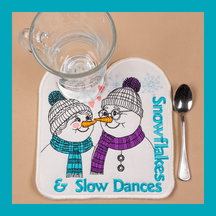 A mug rug with a design of two snowmen wearing hats and scarves, with hearts in the background and the text 'Snowflakes & Slow Dances'.
