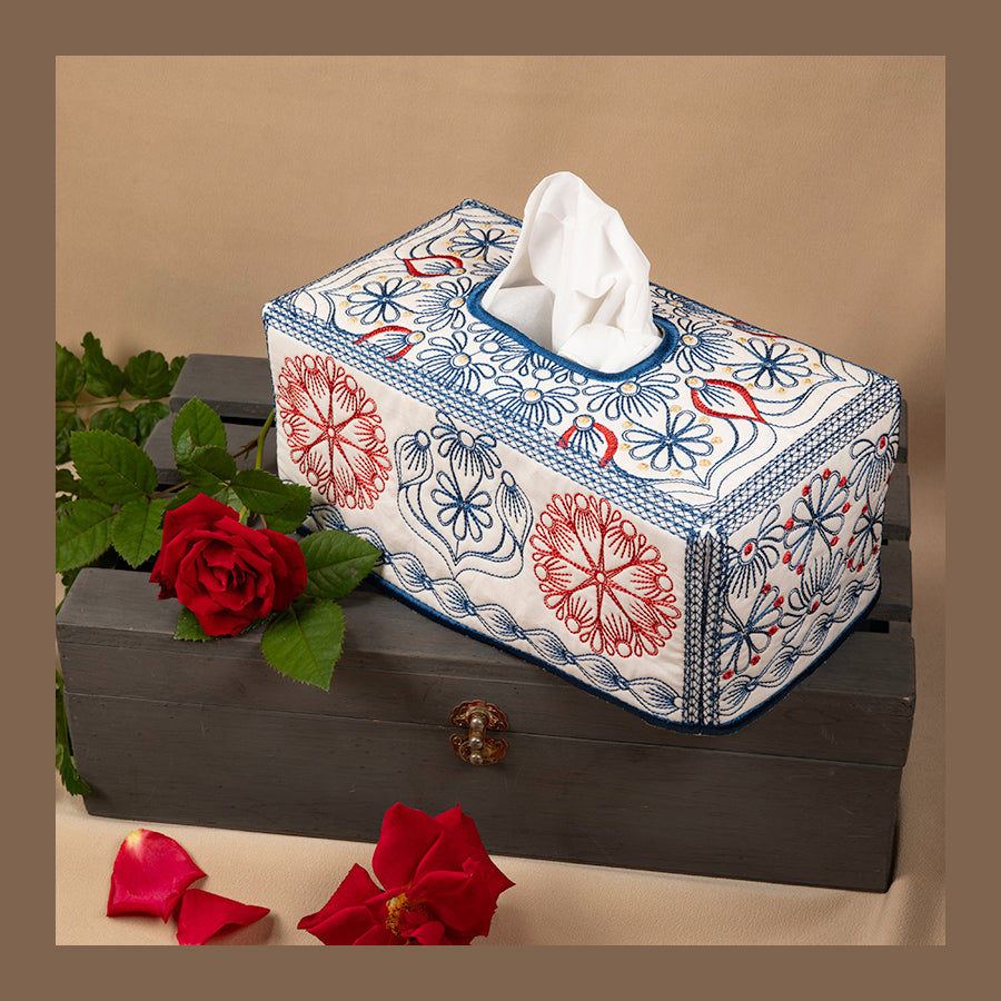 A rectangular tissue box cover with a floral design, featuring a white tissue peeking out from the front, placed on a table with red roses and green leaves around it.