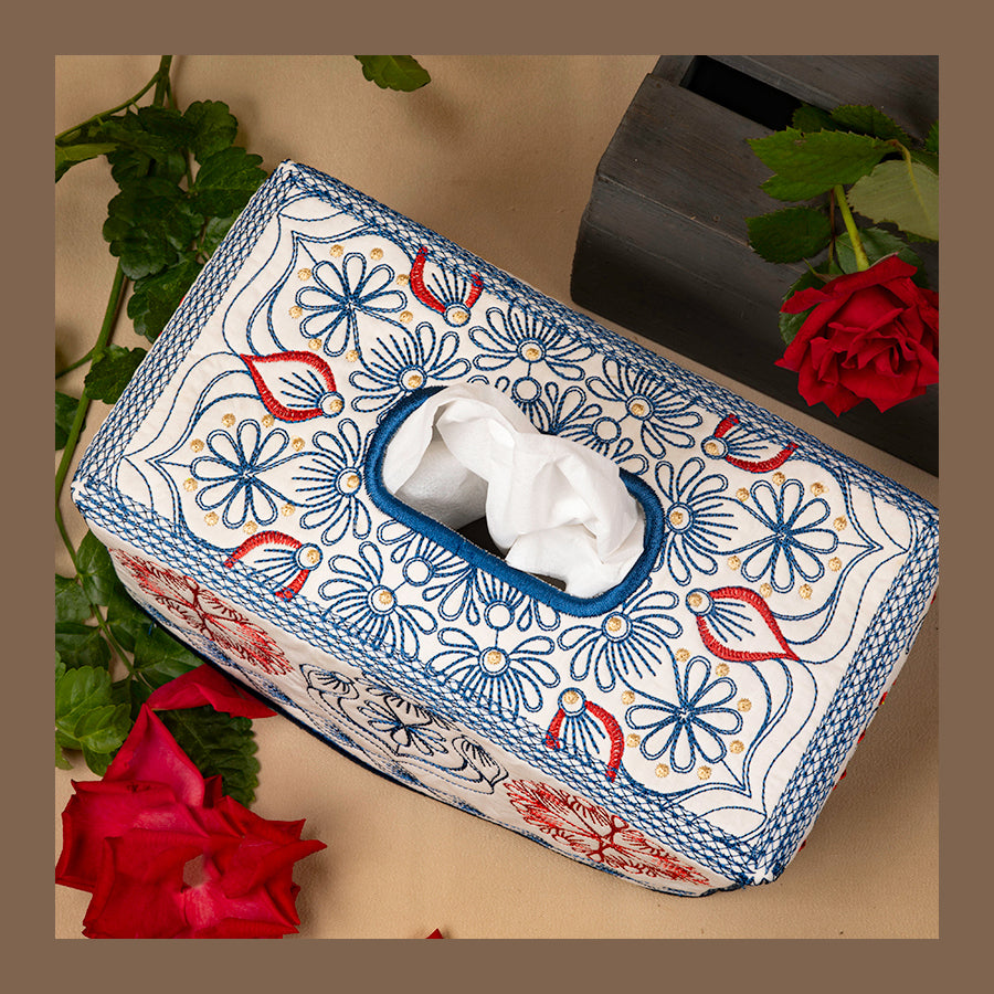 A rectangular tissue box cover with a floral design, featuring a white tissue peeking out from the front, placed on a table with red roses and green leaves around it.