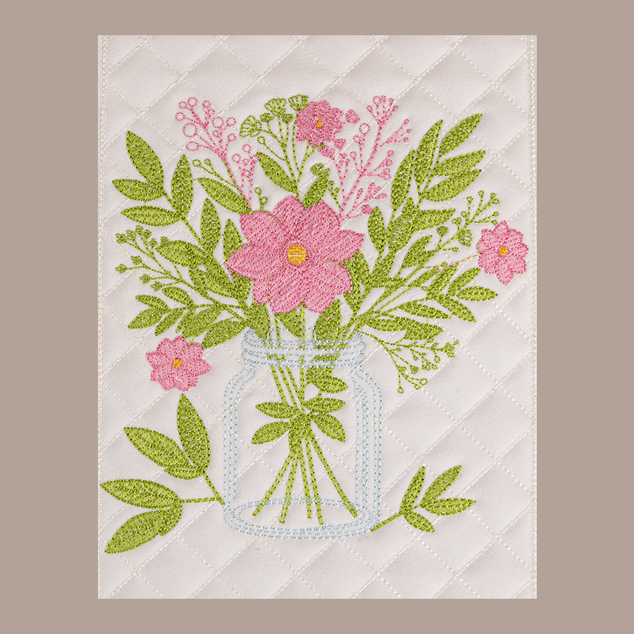 A white quilted placemat with an embroidered floral design in a glass vase, styled on a round wooden table with a folded napkin, a glass of coffee, and a small bouquet of red and white flowers.