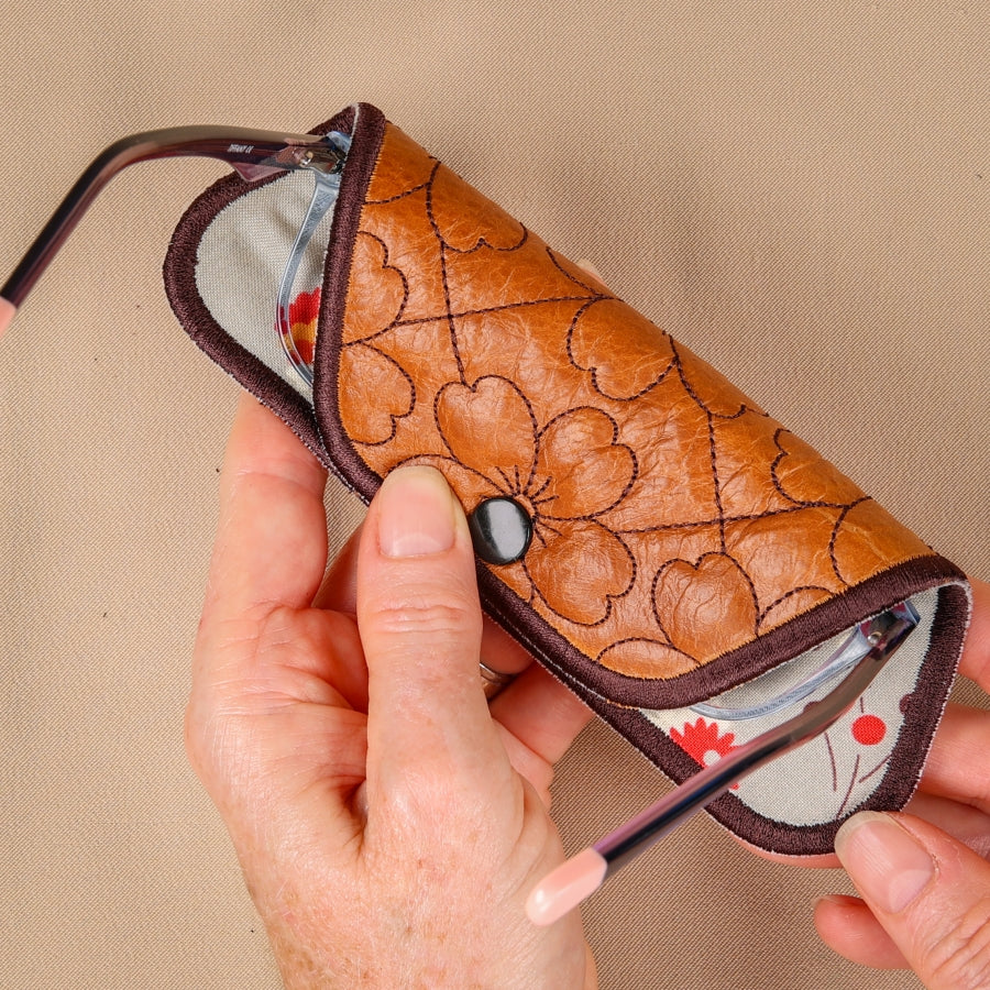 A hand holding a brown eyeglass sleeve with a floral pattern and a blue lens cleaning mechanism visible.