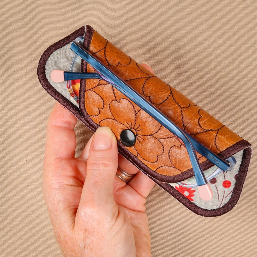 A hand holding a brown eyeglass sleeve with a floral pattern and a blue lens cleaning mechanism visible.