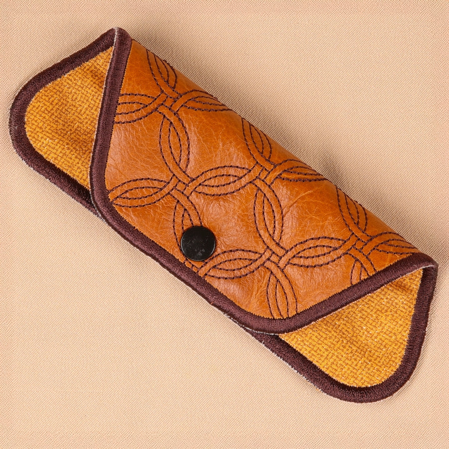 A hand holding a brown eyeglass sleeve with a geometric pattern. The sleeve is designed to hold glasses and has a lens cleaner built into the lining.