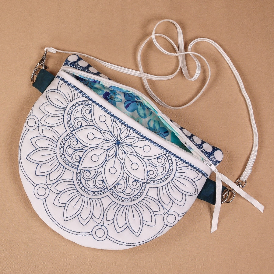 A hand-stitched zipper bag with a mandala design in blue and white colors, with a white zipper and a white shoulder strap on a beige background.