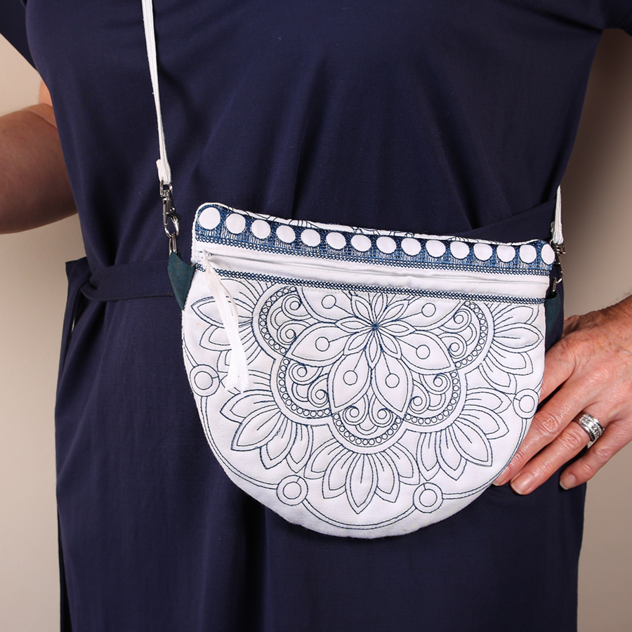 A hand-stitched zipper bag with a mandala design in blue and white colors, with a white zipper and a white shoulder strap on a beige background.