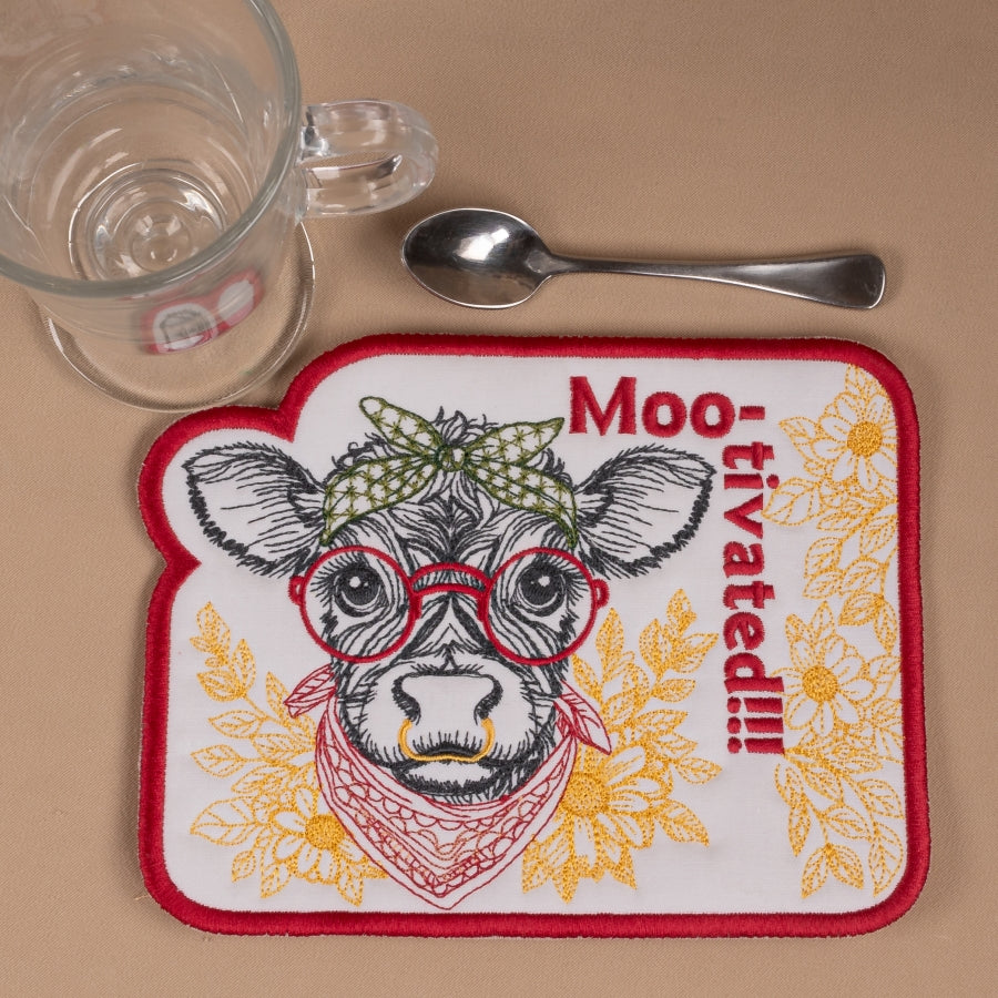 Coaster with cow illustration and 'Moo-tivated!' text on a beige surface with a glass, spoon, and coaster.