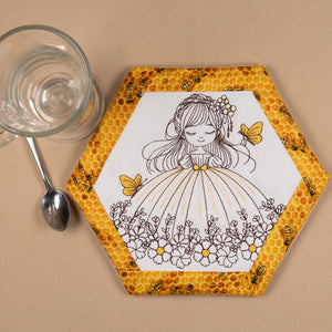 Hexagonal coaster with a design of a girl in a dress surrounded by butterflies and flowers, placed on a beige surface with a glass and spoon.