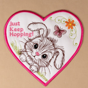 Heart-shaped patch with a kitten illustration and 'Just Keep Hopping!' text on a beige background