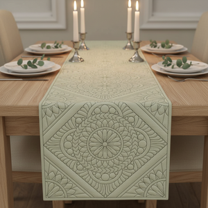 A light sage green table runner with an embossed geometric and floral mandala pattern, displayed on a wooden dining table set with plates, candles, and eucalyptus leaves.