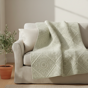 A light sage green quilted throw blanket with intricate mandala and geometric patterns draped over a beige fabric sofa in a sunlit room, next to a potted olive tree.
