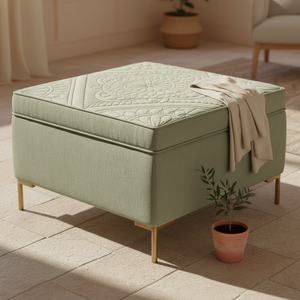 A square ottoman in a light sage green fabric with a quilted top pattern, supported by slim gold-toned legs, styled in a sunlit room with a small potted plant and a draped throw blanket.