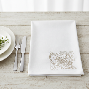 A folded white linen napkin with a delicate beige paisley embroidery in the lower right corner, styled on a light wood tabletop next to a plate, fork, and knife.