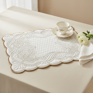 A white quilted table runner with scalloped edges and floral embroidery, styled on a cream tablecloth with a teacup, saucer, flowers, and a white napkin.
