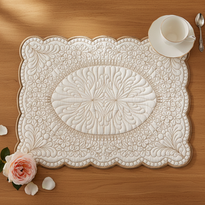An off-white quilted placemat with an intricate floral pattern and scalloped edges, styled on a wooden table with a white teacup, a spoon, and a pink rose. by astitchahalf.net