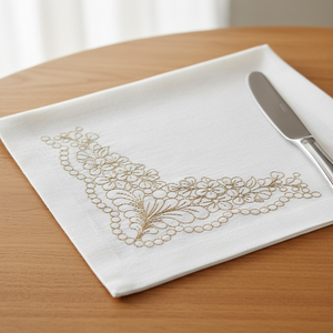 A white linen napkin with delicate gold floral embroidery in the corner, folded on a wooden table next to a silver butter knife. by astitchahalf.net