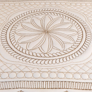 Close-up of a cream-colored fabric with detailed tan embroidery featuring a central floral mandala design and concentric circular patterns.