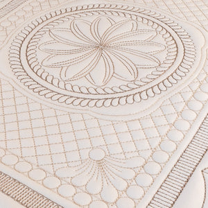 A close-up of a cream-colored quilted fabric with intricate beige embroidery, featuring a central floral motif, a rope border, and a grid pattern.