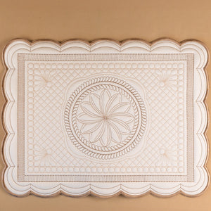 An off-white rectangular decorative mat with a scalloped border and embossed floral medallion pattern, displayed against a solid tan background.