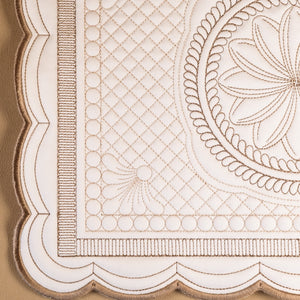 Close-up of an embroidered textile corner showing a cream fabric base with tan embroidery, including a scalloped border, stitched diamond pattern, and floral medallion detail.