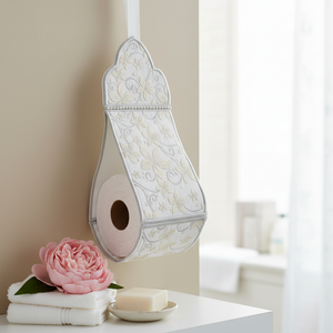 An ornate white toilet paper holder with a floral pattern hangs on a bathroom wall above a white vanity table. A pink rose, folded white towels, and a bar of soap are arranged below, with a softly blurred bathroom background.