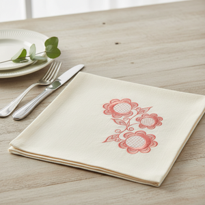 A folded cream-colored linen napkin with a pink embroidered floral pattern, styled on a light wood tabletop next to a silver knife, fork, and white plates with green leaves by astitchahalf.net