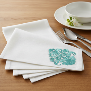 A stack of white linen napkins on a wooden table, each printed with a teal damask pattern in the corner, styled with silver flatware and a white ceramic bowl in the background by astitchahalf.net