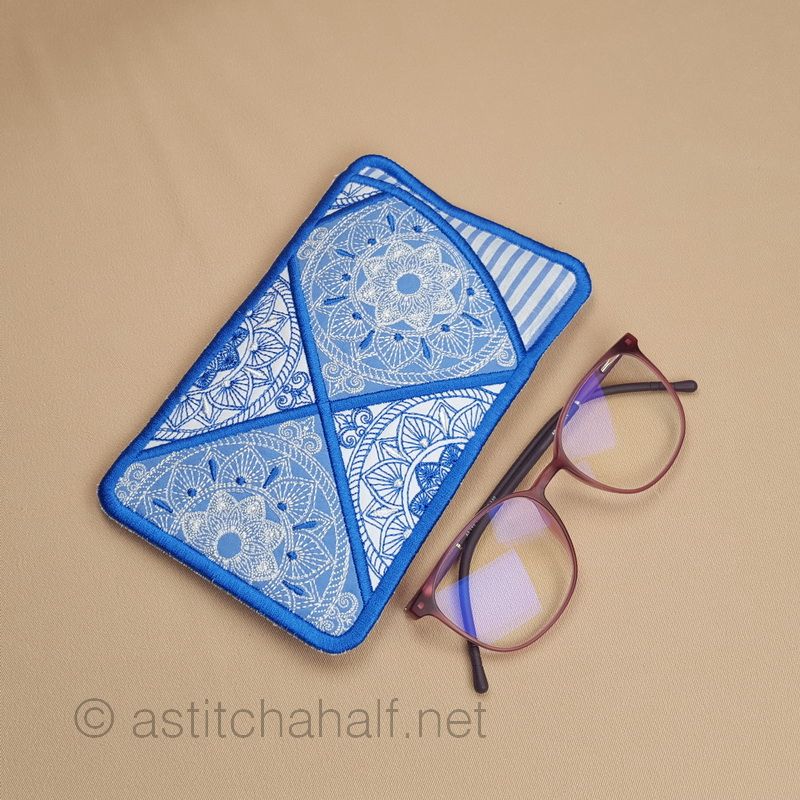 An embroidered eyeglass case with blue binding featuring geometric mandala-style patterns in white stitching on blue fabric sections. A pair of brown-framed glasses with purple-tinted lenses is positioned beside it.
