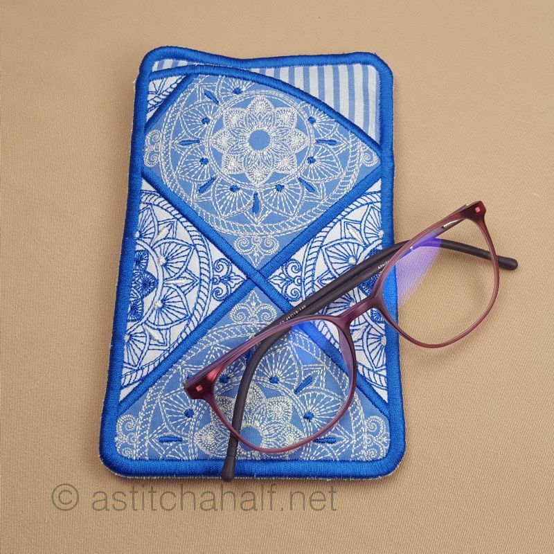 An embroidered eyeglass case with blue binding featuring geometric mandala-style patterns in white stitching on blue fabric sections. A pair of brown-framed glasses with purple-tinted lenses is positioned beside it.