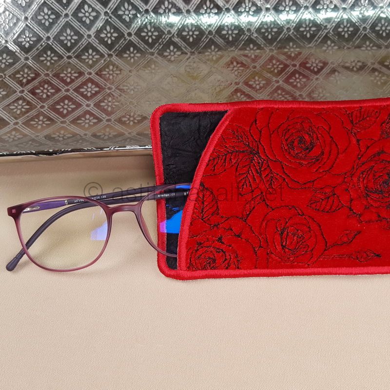 A red eyeglass case with a floral pattern displayed next to a pair of glasses, placed on a textured silver surface.
