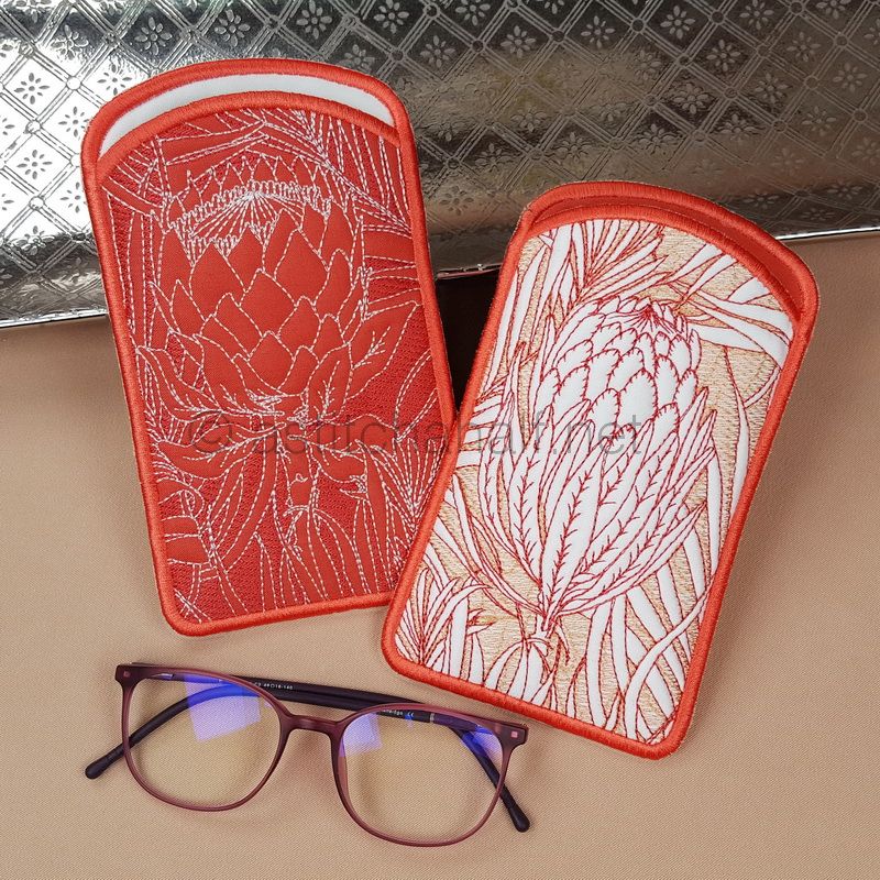 Two embroidered eyeglass cases with red binding, featuring protea flower designs - one on red fabric and one on cream fabric. A pair of brown-framed glasses with blue-tinted lenses is positioned below them.