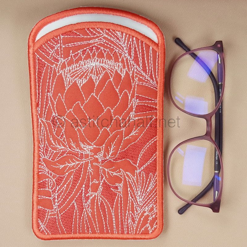 Two embroidered eyeglass cases with red binding, featuring protea flower designs - one on red fabric and one on cream fabric. A pair of brown-framed glasses with blue-tinted lenses is positioned below them.