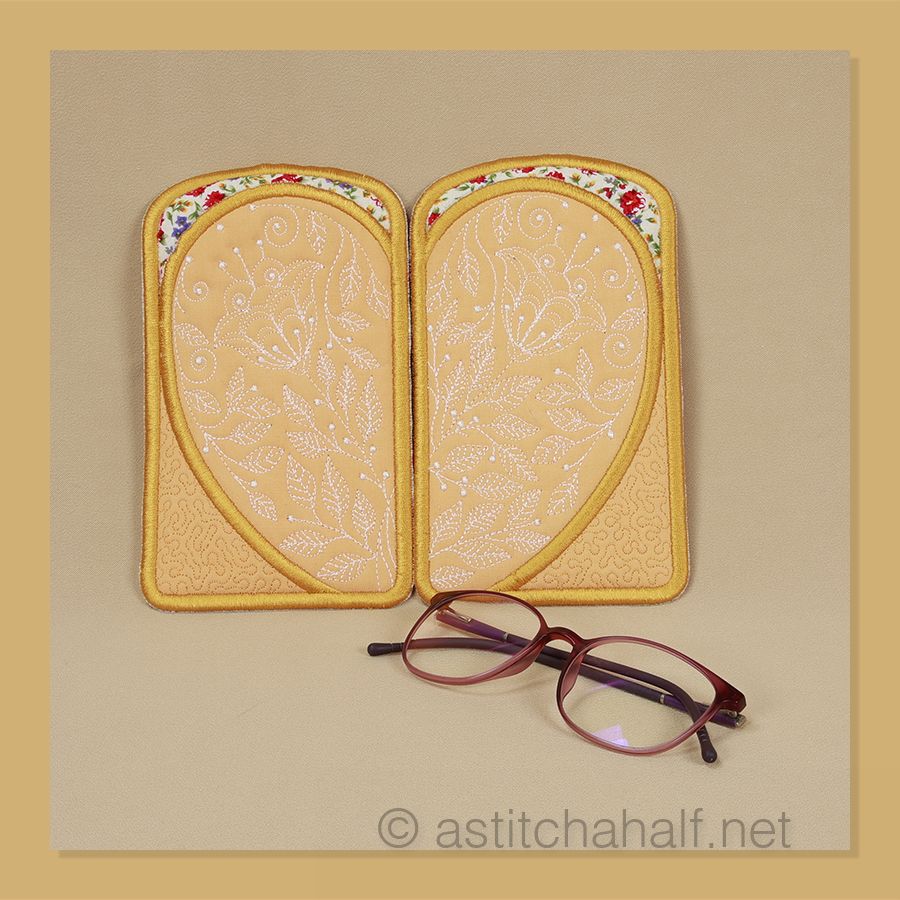 I can see two decorative eyeglass cases with a pair of glasses displayed below them. The cases appear to be handmade with fabric covers and golden-yellow binding or trim around the edges.
The left case features a delicate floral pattern with small red, blue, and purple flowers scattered across a light background. The right case has a more subtle design - it appears to be a light peachy or tan color with an embossed or embroidered botanical pattern that includes what looks like flowering stems or branches.