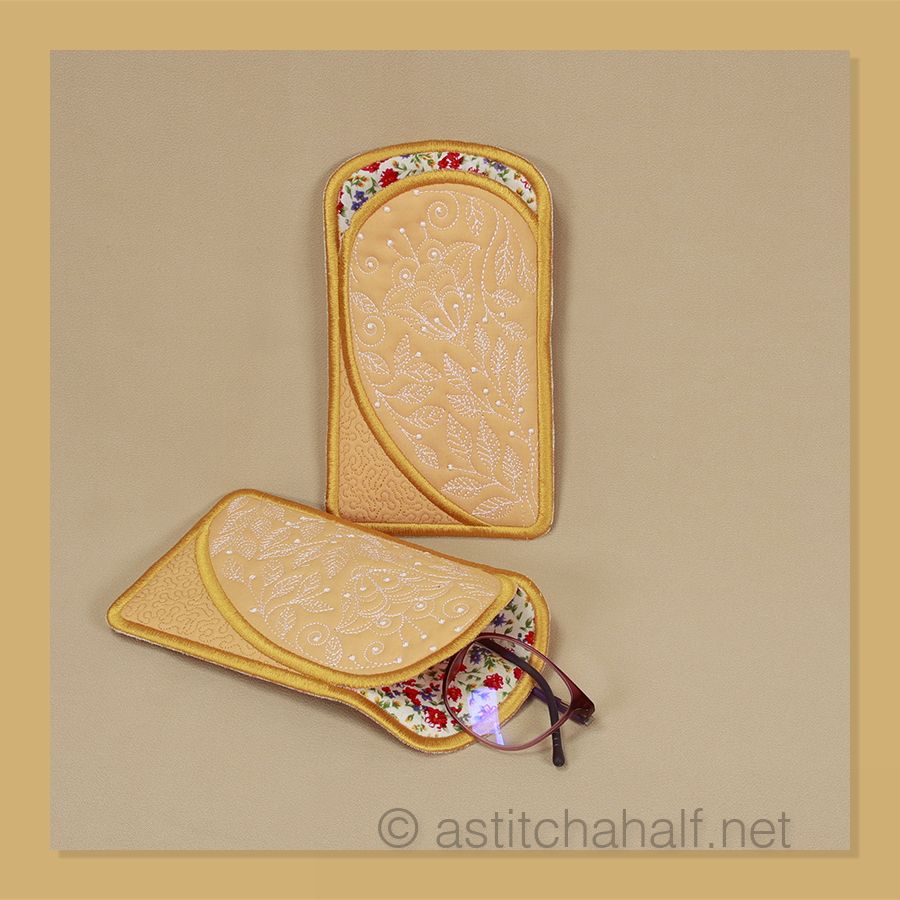 I can see two decorative eyeglass cases with a pair of glasses displayed below them. The cases appear to be handmade with fabric covers and golden-yellow binding or trim around the edges.
The left case features a delicate floral pattern with small red, blue, and purple flowers scattered across a light background. The right case has a more subtle design - it appears to be a light peachy or tan color with an embossed or embroidered botanical pattern that includes what looks like flowering stems or branches.