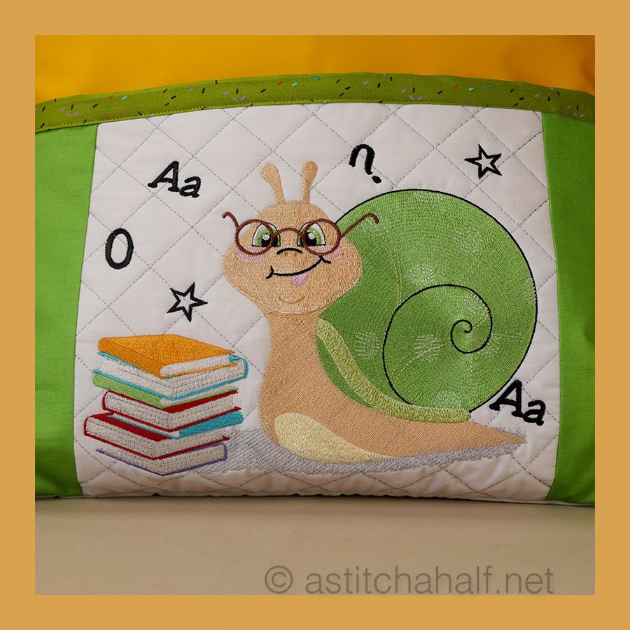 A reading pillow featuring an embroidered design of a snail with glasses, carrying a stack of colorful books, set against a quilted background with letters and stars.