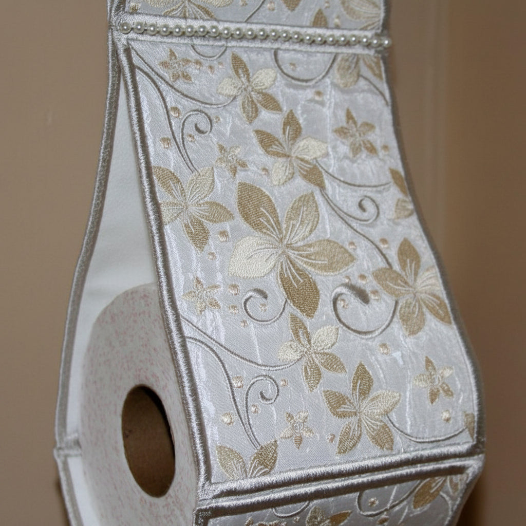 An ornate white toilet paper holder with a floral pattern hangs on a bathroom wall above a white vanity table. A pink rose, folded white towels, and a bar of soap are arranged below, with a softly blurred bathroom background.